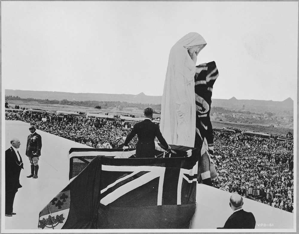 Photographie en noir et blanc – Le roi Édouard III en smoking, dos au photographe, manie des cordes pour dévoiler Mère Canada, couverte de drapeaux (Union Jack et Red Ensign). Une grande foule assiste à la scène de l’inauguration.